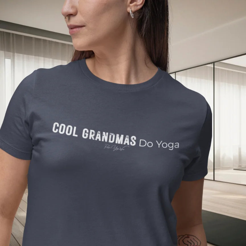 A person with dark hair standing in a bright, airy yoga studio with large windows, wearing the Heather Midnight Navy Grandmas T-Shirt that says Cool Grandmas Do Yoga.