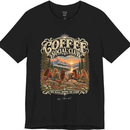 A short sleeved tee called the Grounds T-Shirt, flat lay and a Bella+Canvas 3001 shirt. Simply says, Coffee Social Club, One Ritual Before The Other. Design highlights a group of friends sharing coffee on a rocky mountain outcrop at sunrise, with rolled yoga mats, wildflowers, pine trees and a sleeping dog. Color is Black.