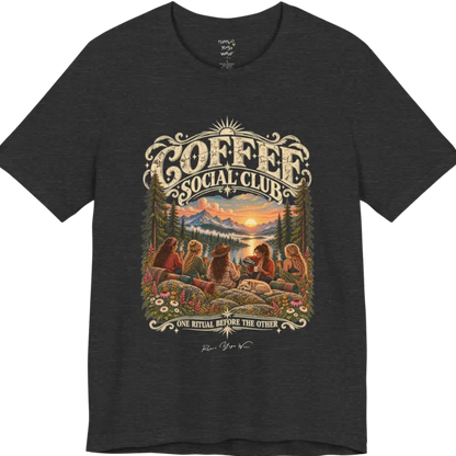 A short sleeved tee called the Grounds T-Shirt, flat lay and a Bella+Canvas 3001 shirt. Simply says, Coffee Social Club, One Ritual Before The Other. Design highlights a group of friends sharing coffee on a rocky mountain outcrop at sunrise, with rolled yoga mats, wildflowers, pine trees and a sleeping dog. Color is Dark Grey Heather.