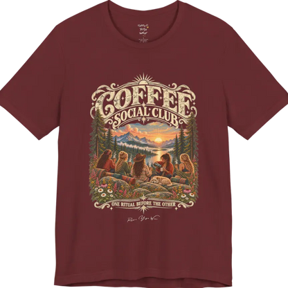 A short sleeved tee called the Grounds T-Shirt, flat lay and a Bella+Canvas 3001 shirt. Simply says, Coffee Social Club, One Ritual Before The Other. Design highlights a group of friends sharing coffee on a rocky mountain outcrop at sunrise, with rolled yoga mats, wildflowers, pine trees and a sleeping dog. Color is Heather Cardinal.