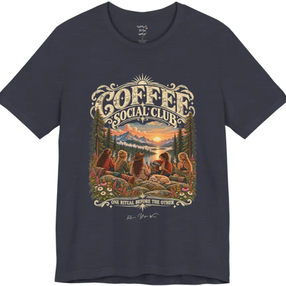 A short sleeved tee called the Grounds T-Shirt, flat lay and a Bella+Canvas 3001 shirt. Simply says, Coffee Social Club, One Ritual Before The Other. Design highlights a group of friends sharing coffee on a rocky mountain outcrop at sunrise, with rolled yoga mats, wildflowers, pine trees and a sleeping dog. Color Heather Midnight Navy.