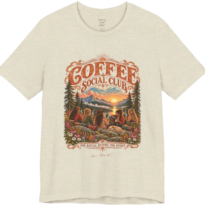 A short sleeved tee called the Grounds T-Shirt, flat lay and a Bella+Canvas 3001 shirt. Simply says, Coffee Social Club, One Ritual Before The Other. Design highlights a group of friends sharing coffee on a rocky mountain outcrop at sunrise, with rolled yoga mats, wildflowers, pine trees and a sleeping dog. Color Heather Prism Natural.