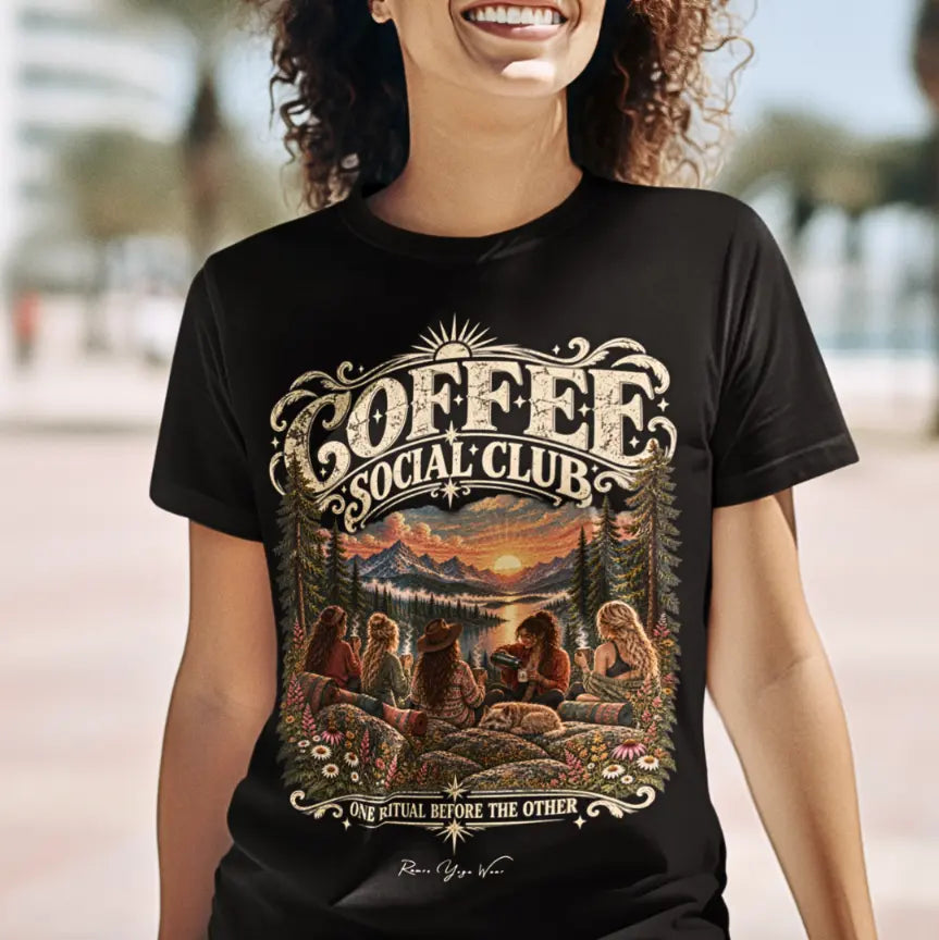 A smiling person with curly hair wearing the black Grounds T-Shirt outdoors, with the Coffee Social Club mountain sunrise design printed across the chest. Color is Black.