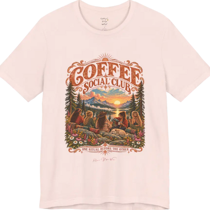 A short sleeved tee called the Grounds T-Shirt, flat lay and a Bella+Canvas 3001 shirt. Simply says, Coffee Social Club, One Ritual Before The Other. Design highlights a group of friends sharing coffee on a rocky mountain outcrop at sunrise, with rolled yoga mats, wildflowers, pine trees and a sleeping dog. Color is Soft Pink.