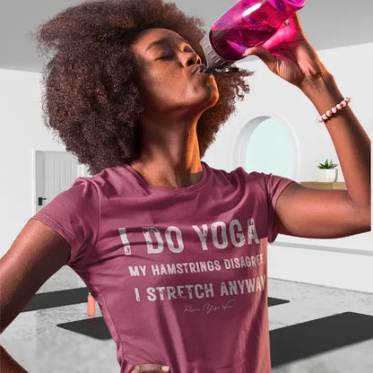 A woman with curly dark hair drinking from a pink water bottle in a bright yoga studio, wearing the Hamstrings T-Shirt with the bold stacked typography on the front. Color is Heather Raspberry.