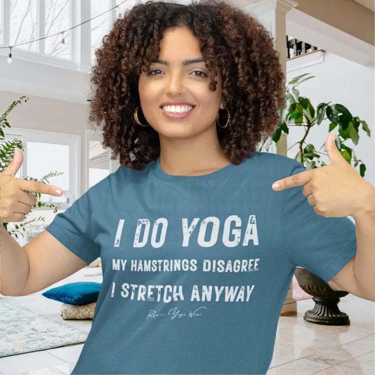 A smiling woman with curly dark hair pointing to the front of her Hamstrings T-Shirt in a bright yoga studio, with the bold stacked typography on the front. Color is Heather Deep Teal.
