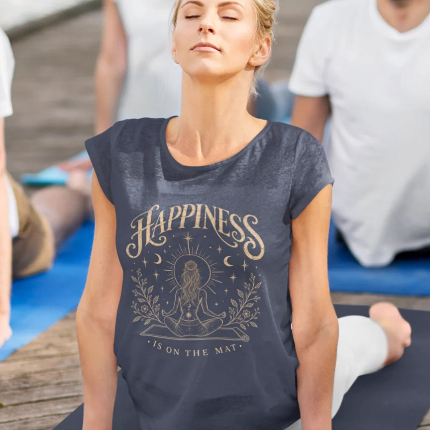 Person with eyes closed and head tilted back in cobra pos wearing a heather navy Happiness T-Shirt with ornate gold lettering and a meditating figure on a yoga mat, at an outdoor yoga class.