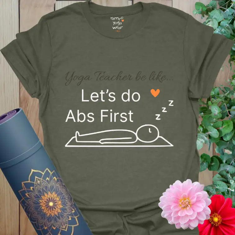 Heather Military Green Abs First T-Shirt with white text reading “Yoga Teacher be like… Let’s do Abs First.” Playful yoga-inspired humor graphic featuring a relaxed figure in Savasana and Ramro’s orange heart for balance and warmth.