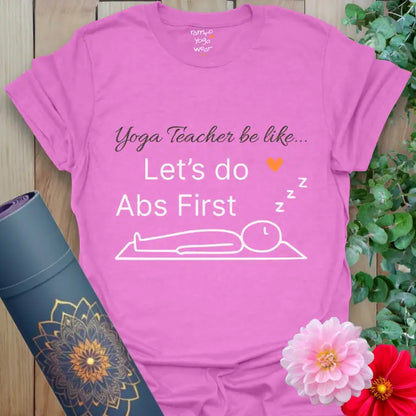 Heather Radiant Orchid Abs First T-Shirt featuring the phrase “Yoga Teacher be like… Let’s do Abs First” in crisp white font. Fun yoga humor design with a peaceful stick figure lying on a mat and an orange heart pop.