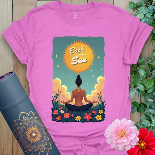 Radiant orchid-colored tee with golden sunrise, seated yogi, and wildflowers beneath “Bask in the Sun” text for soulful living.