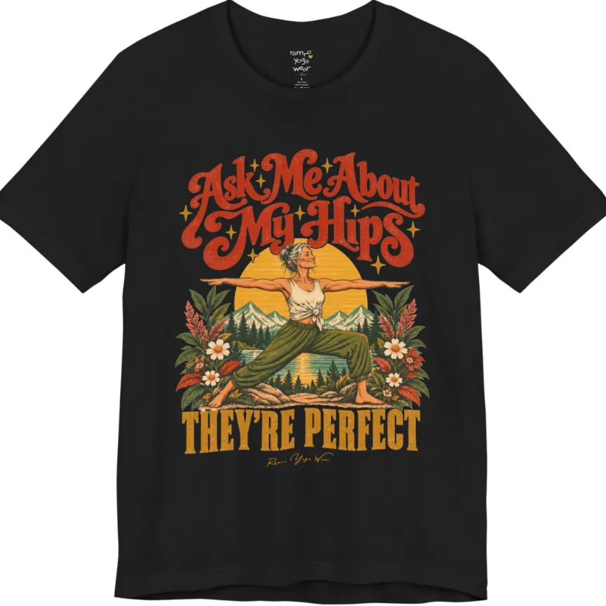A short sleeved tee called the Hips T-Shirt, flat lay and a soft cotton shirt. Simply says, Ask Me About My Hips. They're Perfect. Design highlights a silver-haired woman in warrior two pose, arms outstretched and smiling, in front of a golden sun, mountains, a lake and wildflowers in a retro travel-poster style. Color is  Black.