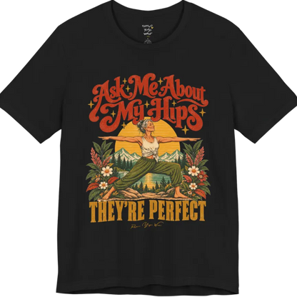 A short sleeved tee called the Hips T-Shirt, flat lay and a soft cotton shirt. Simply says, Ask Me About My Hips. They're Perfect. Design highlights a silver-haired woman in warrior two pose, arms outstretched and smiling, in front of a golden sun, mountains, a lake and wildflowers in a retro travel-poster style. Color is  Black.