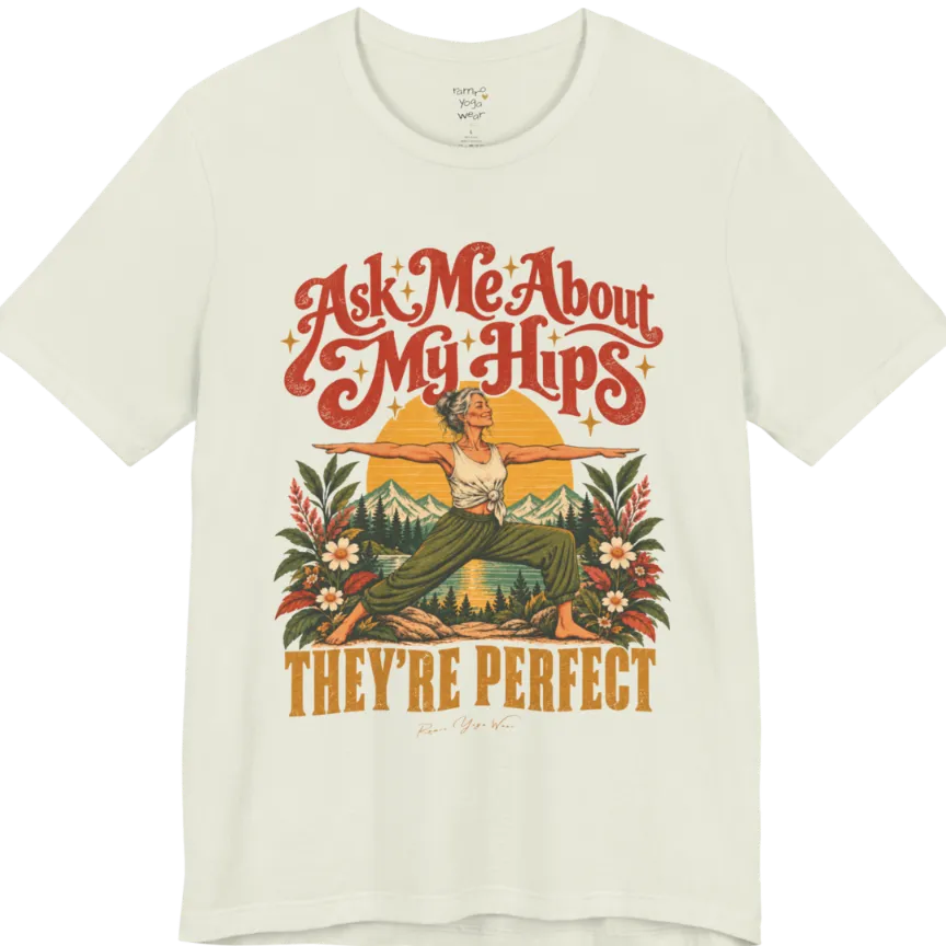 A short sleeved tee called the Hips T-Shirt, flat lay and a soft cotton shirt. Simply says, Ask Me About My Hips. They're Perfect. Design highlights a silver-haired woman in warrior two pose, arms outstretched and smiling, in front of a golden sun, mountains, a lake and wildflowers in a retro travel-poster style. Color is Citron.