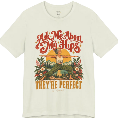 A short sleeved tee called the Hips T-Shirt, flat lay and a soft cotton shirt. Simply says, Ask Me About My Hips. They're Perfect. Design highlights a silver-haired woman in warrior two pose, arms outstretched and smiling, in front of a golden sun, mountains, a lake and wildflowers in a retro travel-poster style. Color is Citron.