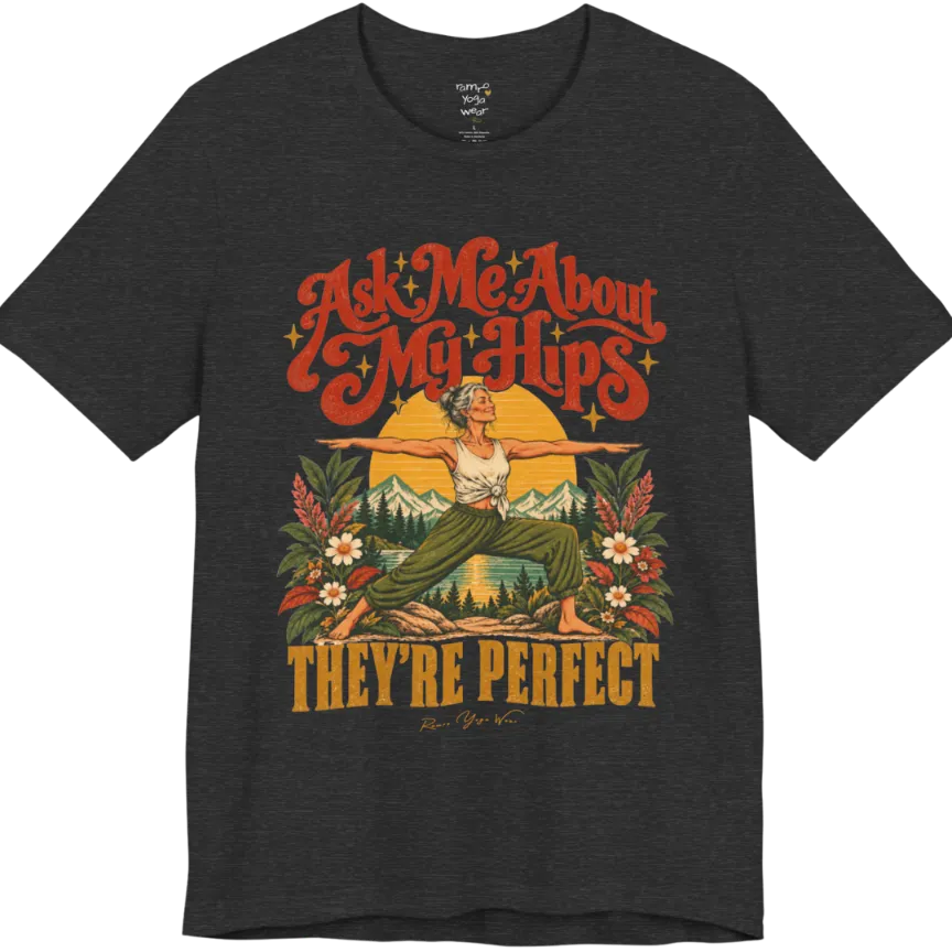 A short sleeved tee called the Hips T-Shirt, flat lay and a soft cotton shirt. Simply says, Ask Me About My Hips. They're Perfect. Design highlights a silver-haired woman in warrior two pose, arms outstretched and smiling, in front of a golden sun, mountains, a lake and wildflowers in a retro travel-poster style. Color is Dark Heather Grey.
