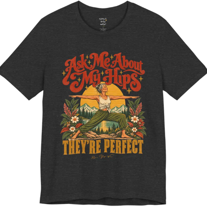 A short sleeved tee called the Hips T-Shirt, flat lay and a soft cotton shirt. Simply says, Ask Me About My Hips. They're Perfect. Design highlights a silver-haired woman in warrior two pose, arms outstretched and smiling, in front of a golden sun, mountains, a lake and wildflowers in a retro travel-poster style. Color is Dark Heather Grey.