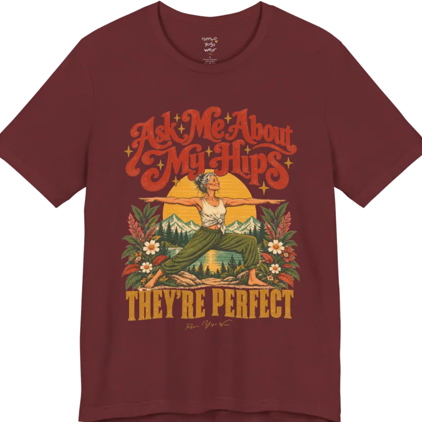 A short sleeved tee called the Hips T-Shirt, flat lay and a soft cotton shirt. Simply says, Ask Me About My Hips. They're Perfect. Design highlights a silver-haired woman in warrior two pose, arms outstretched and smiling, in front of a golden sun, mountains, a lake and wildflowers in a retro travel-poster style. Color is Heather Cardinal.