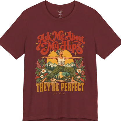 A short sleeved tee called the Hips T-Shirt, flat lay and a soft cotton shirt. Simply says, Ask Me About My Hips. They're Perfect. Design highlights a silver-haired woman in warrior two pose, arms outstretched and smiling, in front of a golden sun, mountains, a lake and wildflowers in a retro travel-poster style. Color is Heather Cardinal.