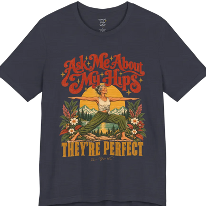 A short sleeved tee called the Hips T-Shirt, flat lay and a soft cotton shirt. Simply says, Ask Me About My Hips. They're Perfect. Design highlights a silver-haired woman in warrior two pose, arms outstretched and smiling, in front of a golden sun, mountains, a lake and wildflowers in a retro travel-poster style. Color is Heather Midnight Navy.