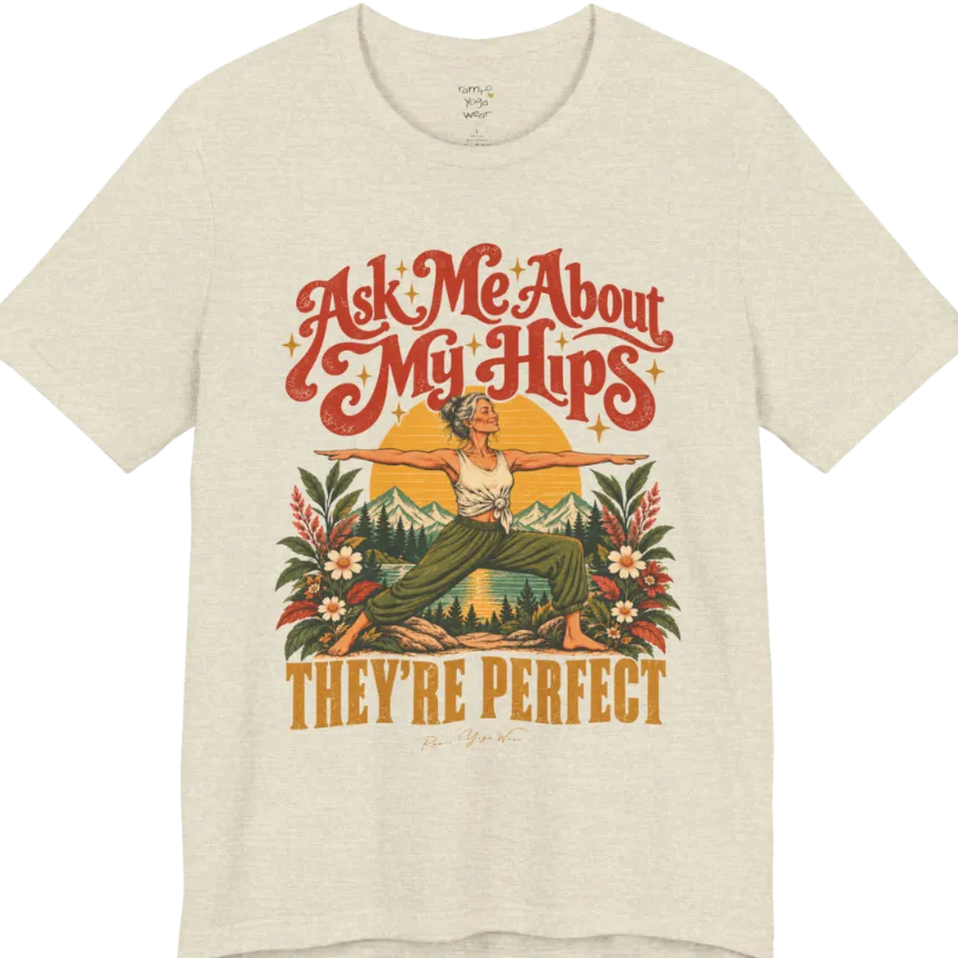 A short sleeved tee called the Hips T-Shirt, flat lay and a soft cotton shirt. Simply says, Ask Me About My Hips. They're Perfect. Design highlights a silver-haired woman in warrior two pose, arms outstretched and smiling, in front of a golden sun, mountains, a lake and wildflowers in a retro travel-poster style. Color is Heather Prism Natural.