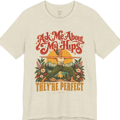 A short sleeved tee called the Hips T-Shirt, flat lay and a soft cotton shirt. Simply says, Ask Me About My Hips. They're Perfect. Design highlights a silver-haired woman in warrior two pose, arms outstretched and smiling, in front of a golden sun, mountains, a lake and wildflowers in a retro travel-poster style. Color is Heather Prism Natural.