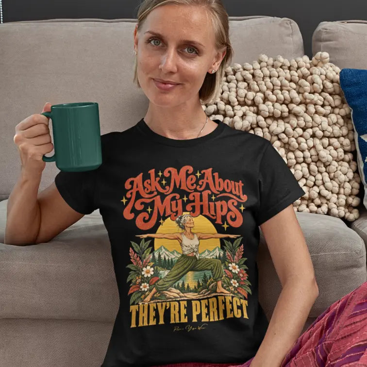 A smiling woman with blonde hair sits next to her sofa holding a green mug, wearing a black Hips T-Shirt with a retro illustration of a woman in warrior two pose.