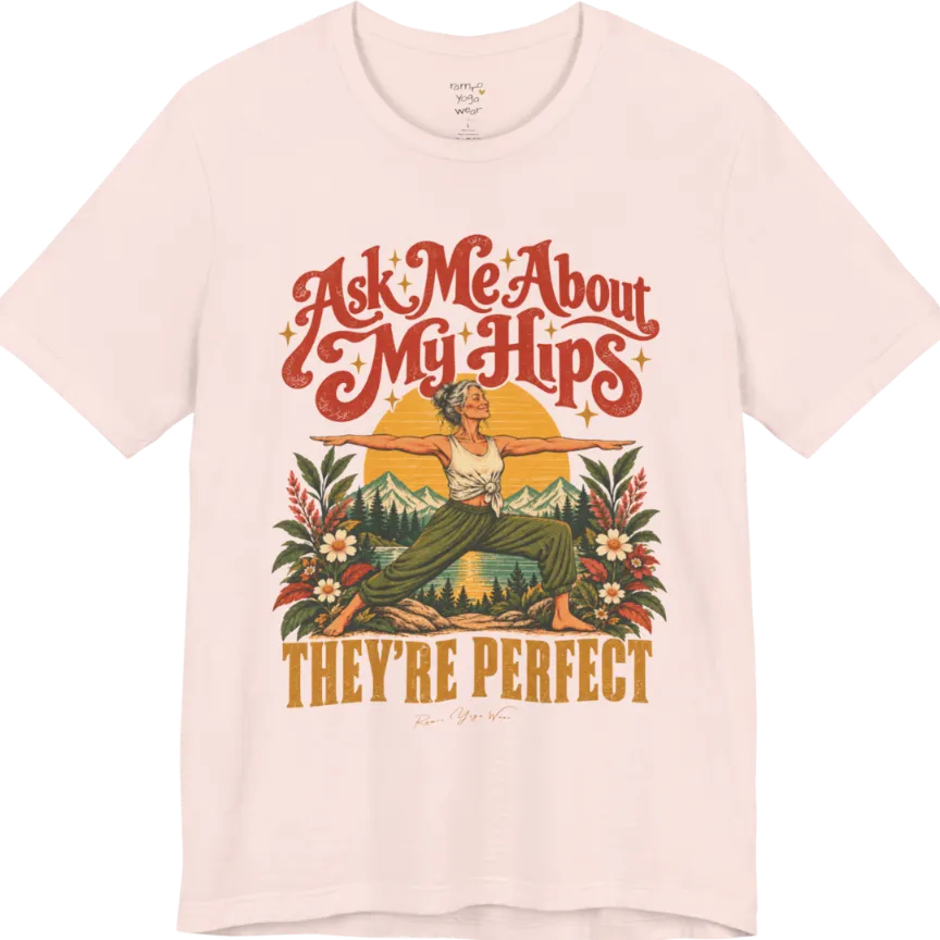 A short sleeved tee called the Hips T-Shirt, flat lay and a soft cotton shirt. Simply says, Ask Me About My Hips. They're Perfect. Design highlights a silver-haired woman in warrior two pose, arms outstretched and smiling, in front of a golden sun, mountains, a lake and wildflowers in a retro travel-poster style. Color is Soft Pink.