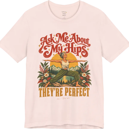 A short sleeved tee called the Hips T-Shirt, flat lay and a soft cotton shirt. Simply says, Ask Me About My Hips. They're Perfect. Design highlights a silver-haired woman in warrior two pose, arms outstretched and smiling, in front of a golden sun, mountains, a lake and wildflowers in a retro travel-poster style. Color is Soft Pink.