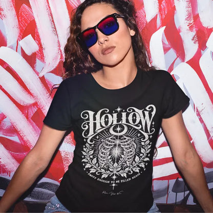 A woman in sunglasses poses in front of a red-and-white painted mural, wearing the Hollow T-Shirt. Hollow T-Shirt is a Gildan 64000 and the color is Black.