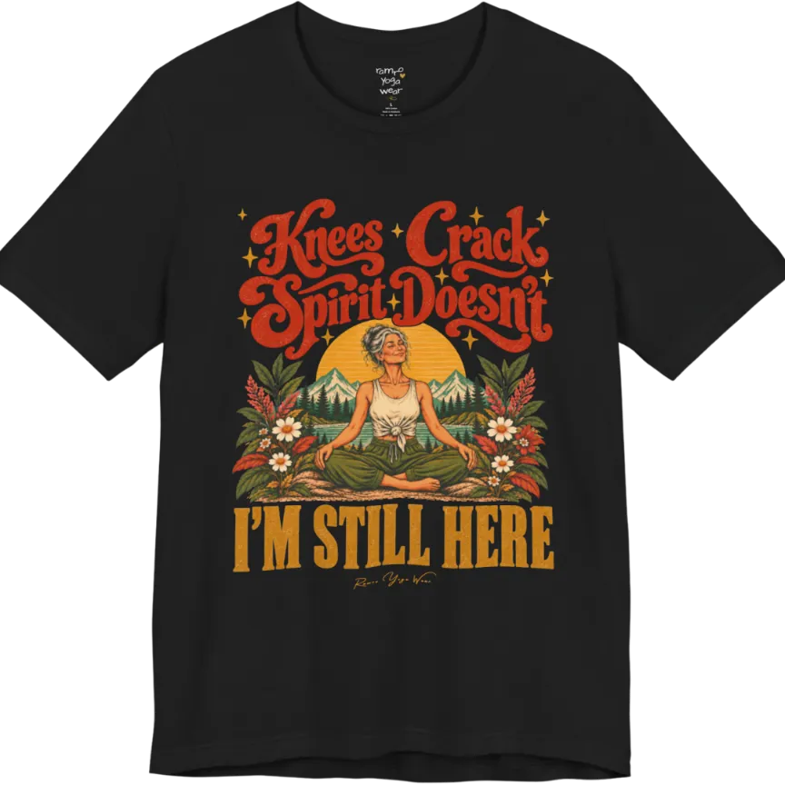 A short sleeved tee called the Knees T-Shirt, flat lay and a soft cotton shirt. Simply says, Knees Crack. Spirit Doesn't. I'm Still Here. Design highlights a silver-haired woman sitting cross-legged with a calm smile, in front of a golden sun, mountains, a lake and wildflowers in a retro travel-poster style. Color is Black.