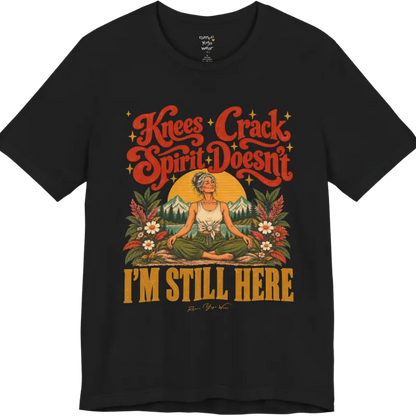 A short sleeved tee called the Knees T-Shirt, flat lay and a soft cotton shirt. Simply says, Knees Crack. Spirit Doesn't. I'm Still Here. Design highlights a silver-haired woman sitting cross-legged with a calm smile, in front of a golden sun, mountains, a lake and wildflowers in a retro travel-poster style. Color is Black.