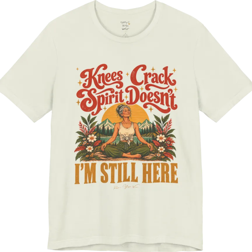 A short sleeved tee called the Knees T-Shirt, flat lay and a soft cotton shirt. Simply says, Knees Crack. Spirit Doesn't. I'm Still Here. Design highlights a silver-haired woman sitting cross-legged with a calm smile, in front of a golden sun, mountains, a lake and wildflowers in a retro travel-poster style. Color is Citron.