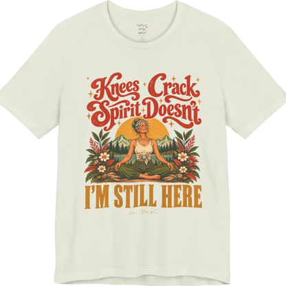 A short sleeved tee called the Knees T-Shirt, flat lay and a soft cotton shirt. Simply says, Knees Crack. Spirit Doesn't. I'm Still Here. Design highlights a silver-haired woman sitting cross-legged with a calm smile, in front of a golden sun, mountains, a lake and wildflowers in a retro travel-poster style. Color is Citron.