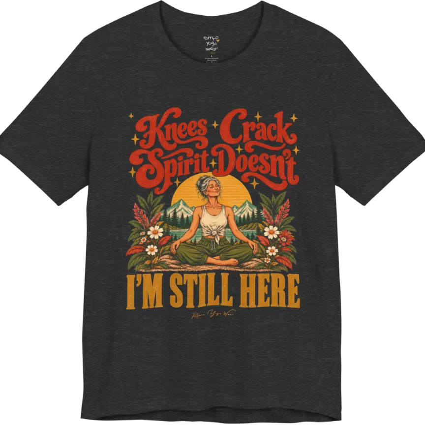 A short sleeved tee called the Knees T-Shirt, flat lay and a soft cotton shirt. Simply says, Knees Crack. Spirit Doesn't. I'm Still Here. Design highlights a silver-haired woman sitting cross-legged with a calm smile, in front of a golden sun, mountains, a lake and wildflowers in a retro travel-poster style. Color is Dark Grey Heather.