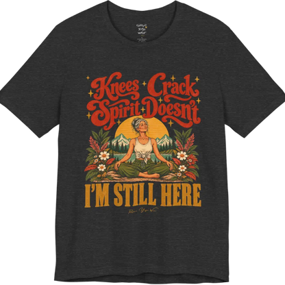 A short sleeved tee called the Knees T-Shirt, flat lay and a soft cotton shirt. Simply says, Knees Crack. Spirit Doesn't. I'm Still Here. Design highlights a silver-haired woman sitting cross-legged with a calm smile, in front of a golden sun, mountains, a lake and wildflowers in a retro travel-poster style. Color is Dark Grey Heather.