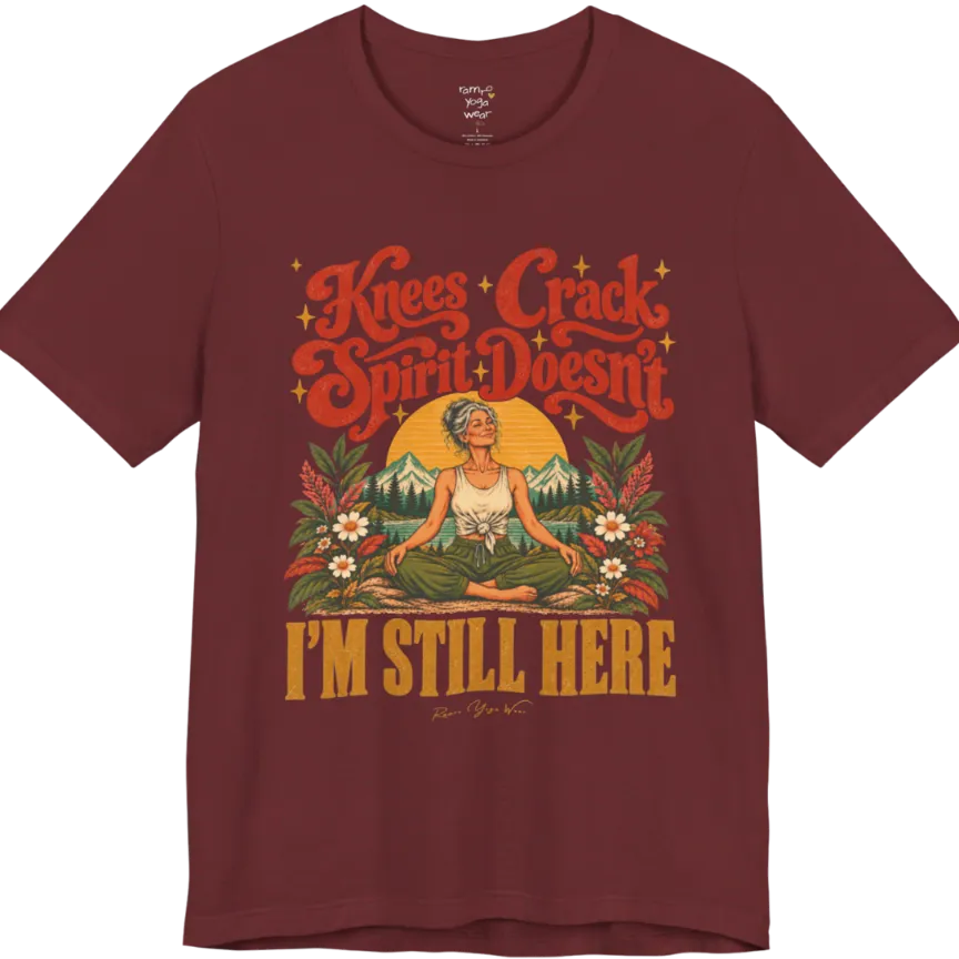 A short sleeved tee called the Knees T-Shirt, flat lay and a soft cotton shirt. Simply says, Knees Crack. Spirit Doesn't. I'm Still Here. Design highlights a silver-haired woman sitting cross-legged with a calm smile, in front of a golden sun, mountains, a lake and wildflowers in a retro travel-poster style. Color is Heather Cardinal.