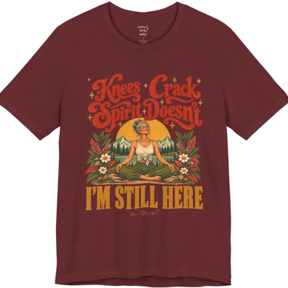 A short sleeved tee called the Knees T-Shirt, flat lay and a soft cotton shirt. Simply says, Knees Crack. Spirit Doesn't. I'm Still Here. Design highlights a silver-haired woman sitting cross-legged with a calm smile, in front of a golden sun, mountains, a lake and wildflowers in a retro travel-poster style. Color is Heather Cardinal.