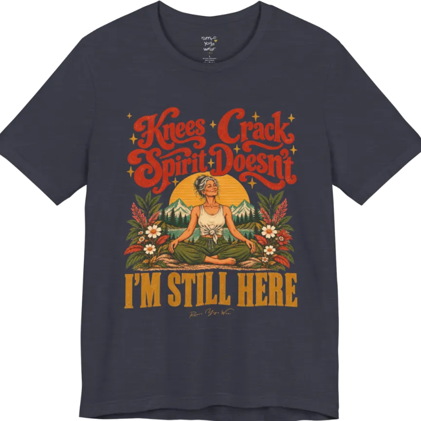 A short sleeved tee called the Knees T-Shirt, flat lay and a soft cotton shirt. Simply says, Knees Crack. Spirit Doesn't. I'm Still Here. Design highlights a silver-haired woman sitting cross-legged with a calm smile, in front of a golden sun, mountains, a lake and wildflowers in a retro travel-poster style. Color is Heather Midnight Navy.