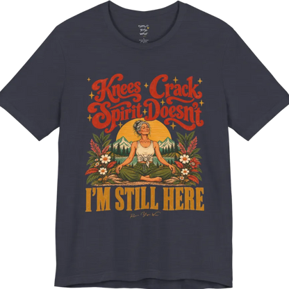 A short sleeved tee called the Knees T-Shirt, flat lay and a soft cotton shirt. Simply says, Knees Crack. Spirit Doesn't. I'm Still Here. Design highlights a silver-haired woman sitting cross-legged with a calm smile, in front of a golden sun, mountains, a lake and wildflowers in a retro travel-poster style. Color is Heather Midnight Navy.
