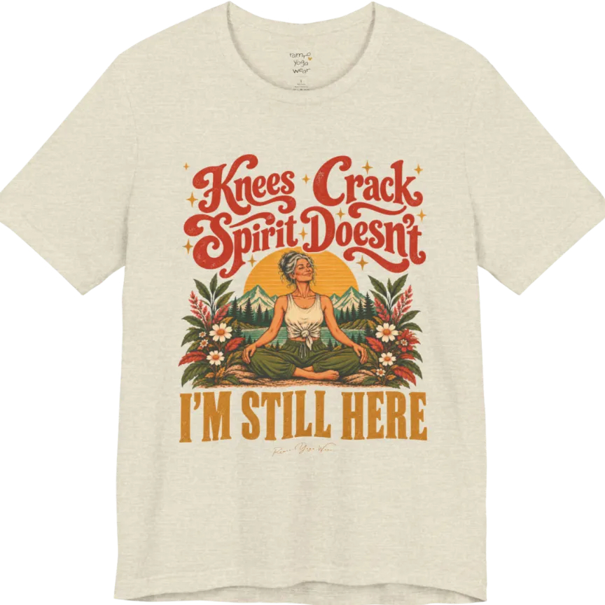 A short sleeved tee called the Knees T-Shirt, flat lay and a soft cotton shirt. Simply says, Knees Crack. Spirit Doesn't. I'm Still Here. Design highlights a silver-haired woman sitting cross-legged with a calm smile, in front of a golden sun, mountains, a lake and wildflowers in a retro travel-poster style. Color is Heather Prism Natural.