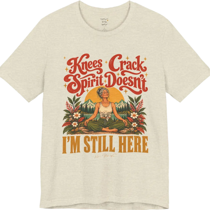 A short sleeved tee called the Knees T-Shirt, flat lay and a soft cotton shirt. Simply says, Knees Crack. Spirit Doesn't. I'm Still Here. Design highlights a silver-haired woman sitting cross-legged with a calm smile, in front of a golden sun, mountains, a lake and wildflowers in a retro travel-poster style. Color is Heather Prism Natural.