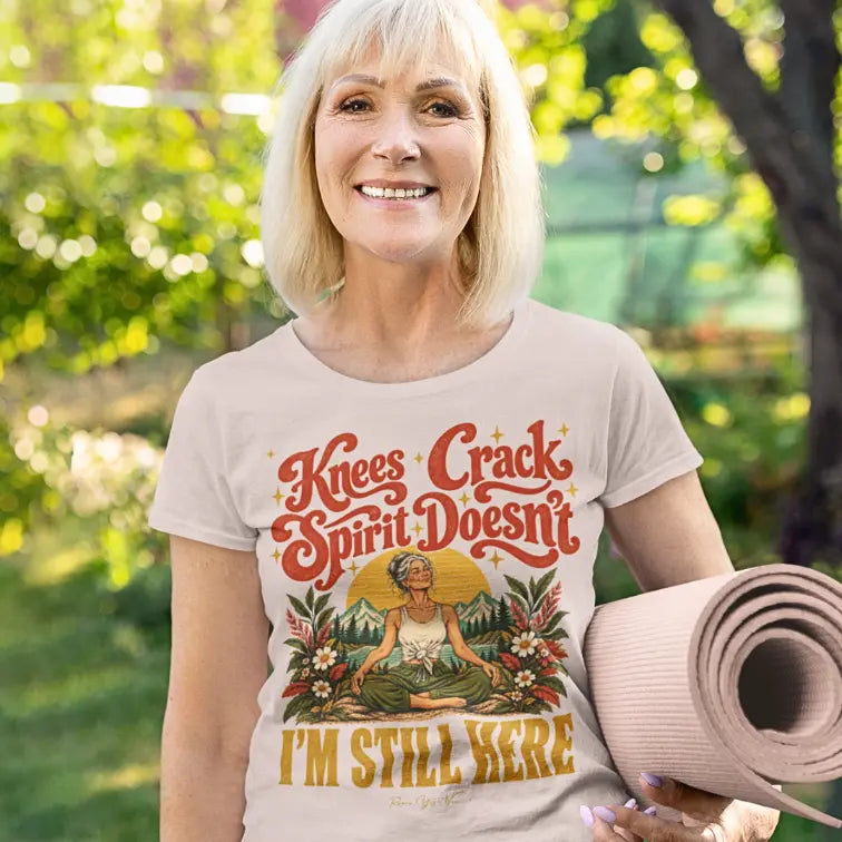 A smiling woman with light blonde hair stands outdoors in a sunlit garden holding a rolled pink yoga mat, wearing a Soft Pink Knees T-Shirt with a retro illustration of a woman sitting cross-legged.