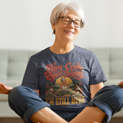 A smiling woman with short grey hair and glasses sits cross-legged on the floor in a bright room, wearing a Heather Midnight Navy Knees T-Shirt with a retro illustration of a woman sitting cross-legged.
