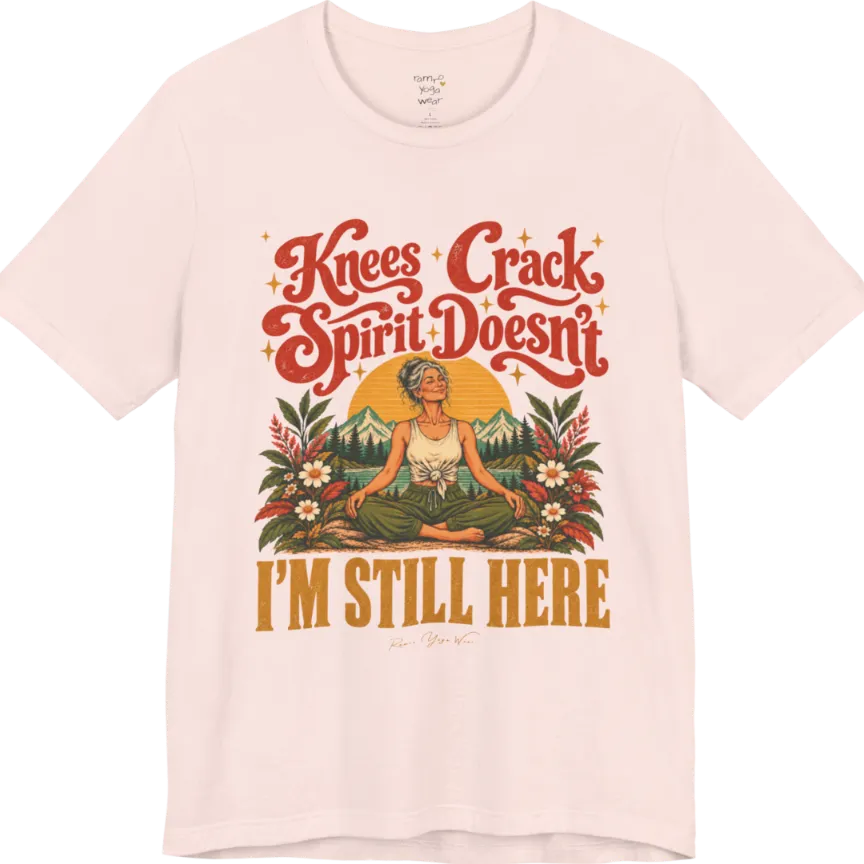 A short sleeved tee called the Knees T-Shirt, flat lay and a soft cotton shirt. Simply says, Knees Crack. Spirit Doesn't. I'm Still Here. Design highlights a silver-haired woman sitting cross-legged with a calm smile, in front of a golden sun, mountains, a lake and wildflowers in a retro travel-poster style. Color is Soft Pink.
