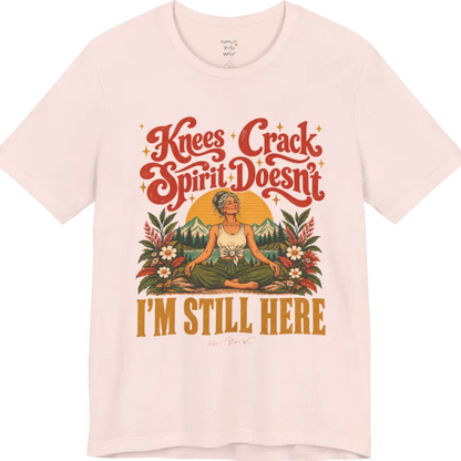 A short sleeved tee called the Knees T-Shirt, flat lay and a soft cotton shirt. Simply says, Knees Crack. Spirit Doesn't. I'm Still Here. Design highlights a silver-haired woman sitting cross-legged with a calm smile, in front of a golden sun, mountains, a lake and wildflowers in a retro travel-poster style. Color is Soft Pink.