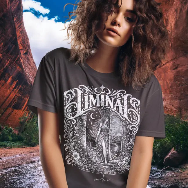 Curly-haired person wearing a dark heather Liminal T-Shirt with ornate white lettering and a figure in an arched doorway, standing in a red rock canyon.