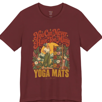 A short sleeved tee called the Mats T-Shirt, flat lay and a soft cotton shirt. Simply says, You Can Never Have Too Many Yoga Mats. Design highlights a silver-haired woman with her hand on her chin, deciding between a wooden rack of rolled yoga mats in rust, mustard, olive, teal and pink, in front of a golden sun, mountains and wildflowers in a retro travel-poster style. Color is Heather Cardinal.