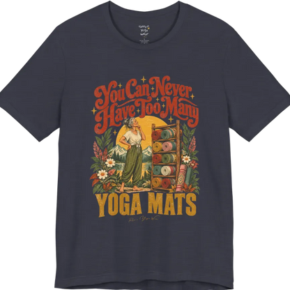 A short sleeved tee called the Mats T-Shirt, flat lay and a soft cotton shirt. Simply says, You Can Never Have Too Many Yoga Mats. Design highlights a silver-haired woman with her hand on her chin, deciding between a wooden rack of rolled yoga mats in rust, mustard, olive, teal and pink, in front of a golden sun, mountains and wildflowers in a retro travel-poster style. Color is Heather Midnight Navy.