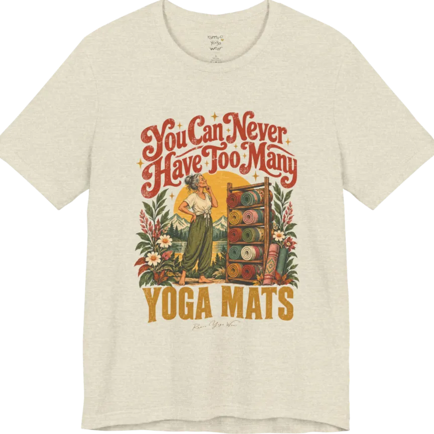 A short sleeved tee called the Mats T-Shirt, flat lay and a soft cotton shirt. Simply says, You Can Never Have Too Many Yoga Mats. Design highlights a silver-haired woman with her hand on her chin, deciding between a wooden rack of rolled yoga mats in rust, mustard, olive, teal and pink, in front of a golden sun, mountains and wildflowers in a retro travel-poster style. Color is Heather Prism Natural.