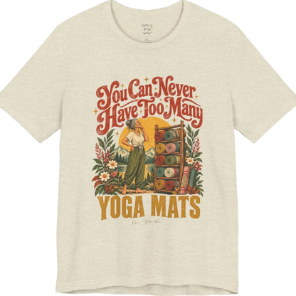 A short sleeved tee called the Mats T-Shirt, flat lay and a soft cotton shirt. Simply says, You Can Never Have Too Many Yoga Mats. Design highlights a silver-haired woman with her hand on her chin, deciding between a wooden rack of rolled yoga mats in rust, mustard, olive, teal and pink, in front of a golden sun, mountains and wildflowers in a retro travel-poster style. Color is Heather Prism Natural.